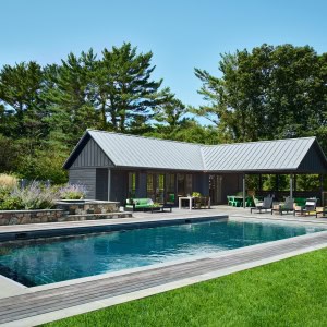 The pool, hot tub, patio, and pool house are in a gated area surrounded by flowers, bushes, and greenery that blend into their surroundings. Like the farmhouse, the pool house has a metal roof and primitive wood siding.