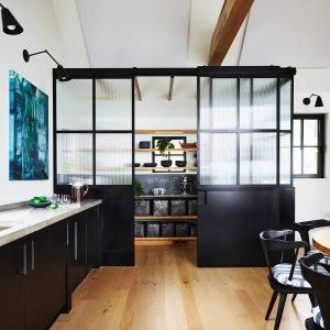 The kitchen pantry is fashioned to mimic an industrial storage room with steel and glass panels encasing surround shelving which are topped off with high windows for natural light.  The pantry's glass panels are not fully transparent, hiding the cans, bowls, and cups stacked inside.